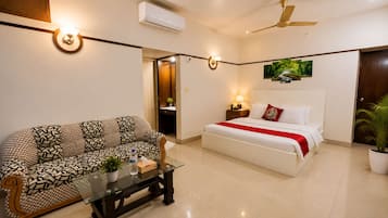 A hotel room with a sofa, a bed with a red blanket, a bedside table with a lamp, and a ceiling fan.