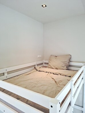 Cama de solteiro com estrutura de madeira e colchão bege em quarto com paredes brancas e luminária no teto.