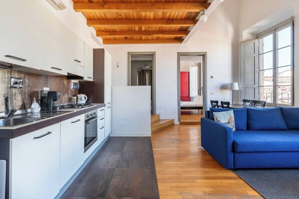 Een moderne keuken met witte kasten en een houten plafond, een zithoek met een blauwe bank en een lamp, en uitzicht op een rode kamer door een geopende deur.