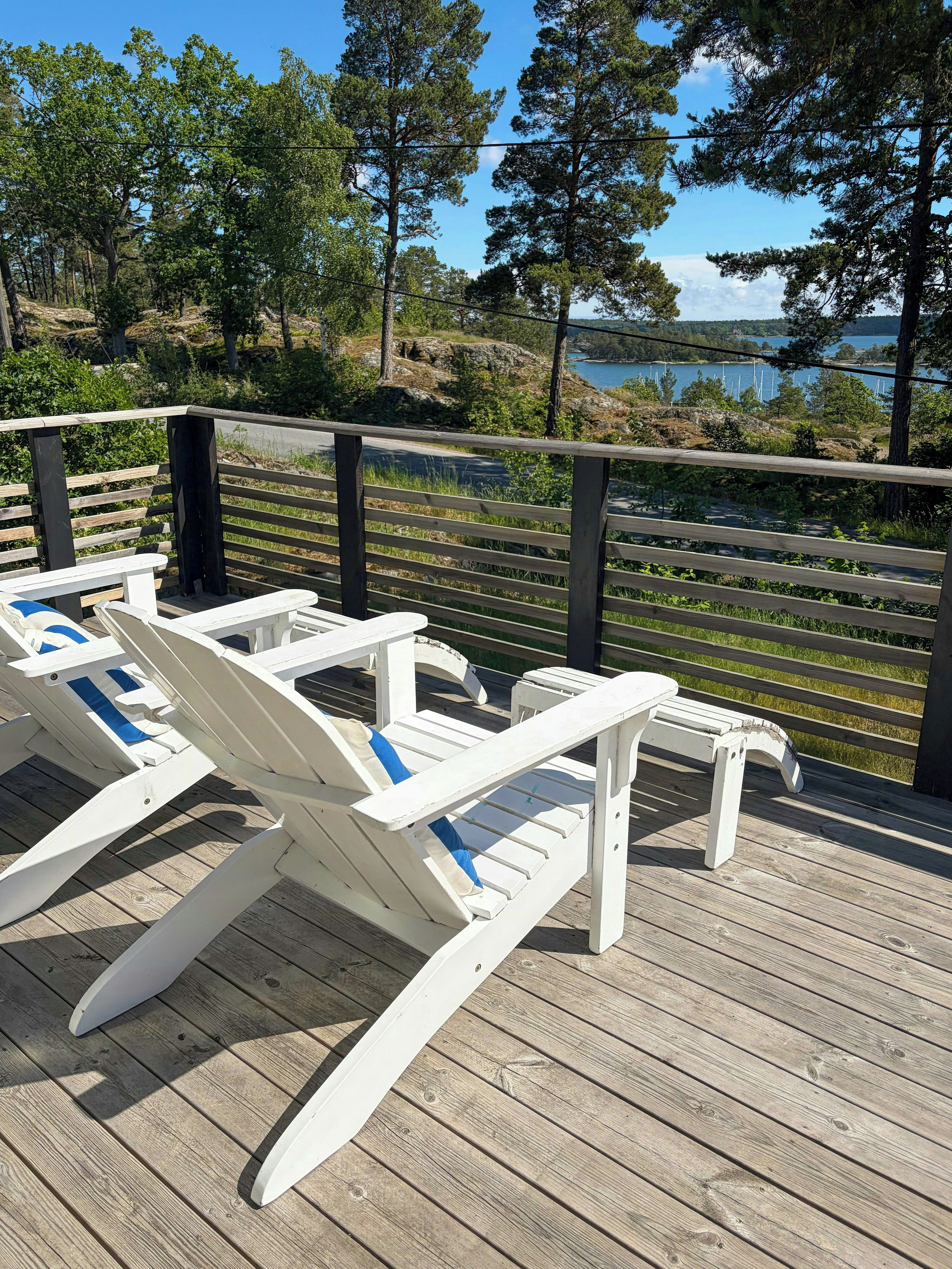 Una terraza de madera con sillas reclinables blancas y una barandilla, con vista a un cuerpo de agua y árboles.