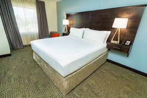 Habitación de hotel con una cama grande, dos mesitas de noche con lámparas, una silla y vistas al exterior a través de cortinas translúcidas.