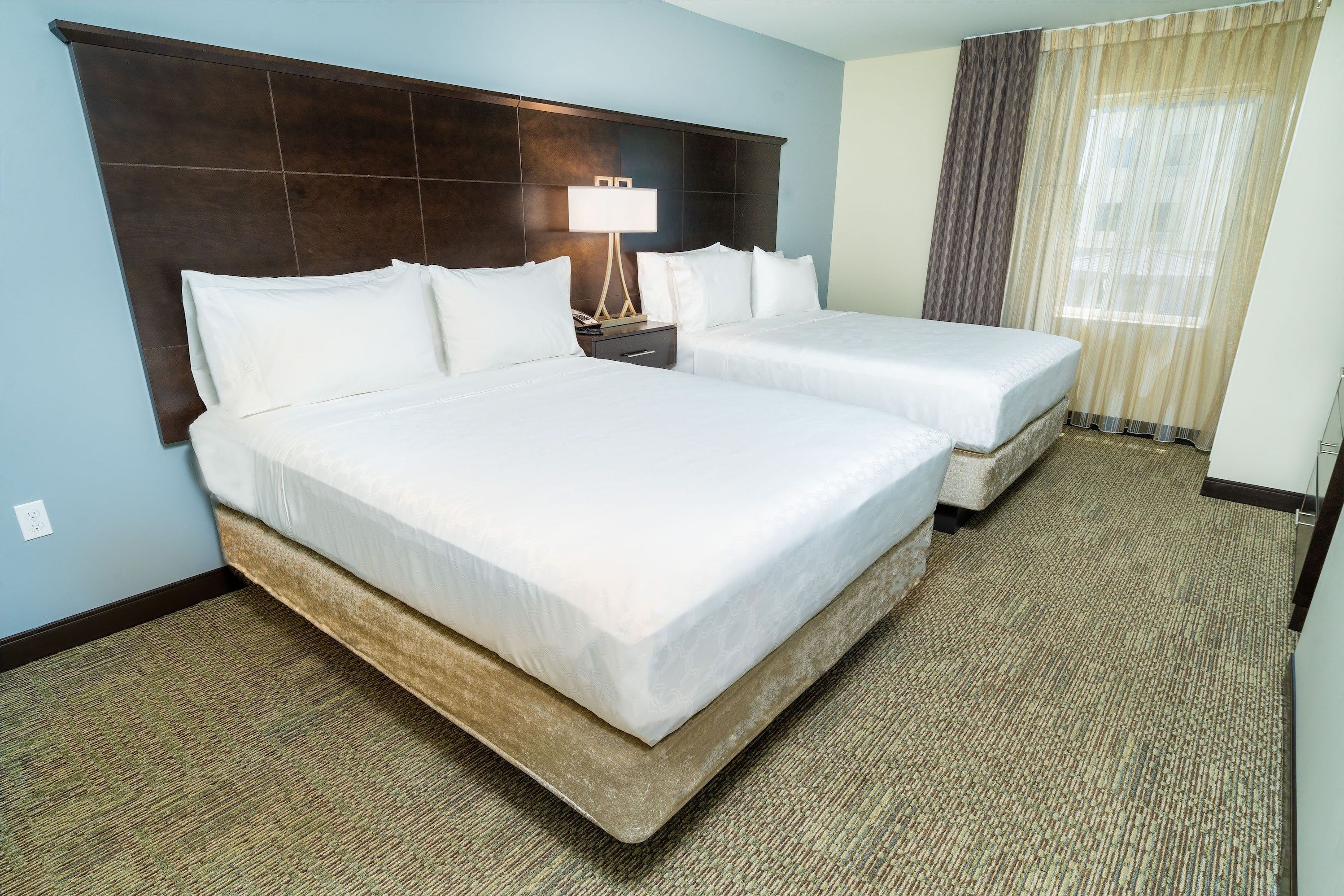 A hotel room with two beds, a dark headboard, a nightstand with a lamp, and a window with curtains.