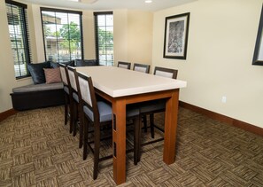 A dining area with a rectangular table, six chairs, a bench with cushions, and large windows with blinds.