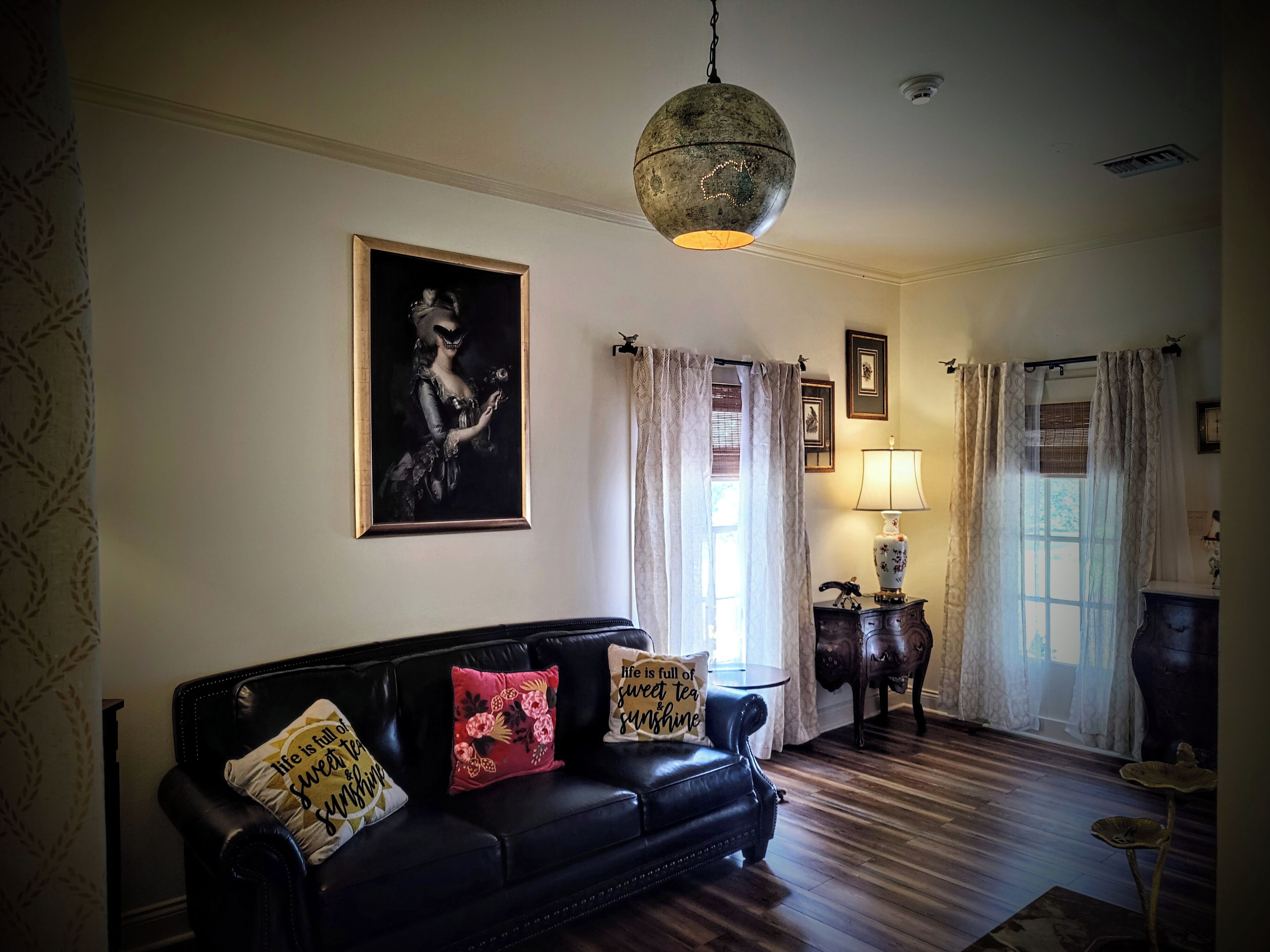 Una sala de estar con un sofá de cuero negro, una lámpara colgante tipo globo, un retrato enmarco y una mesa con lámpara.