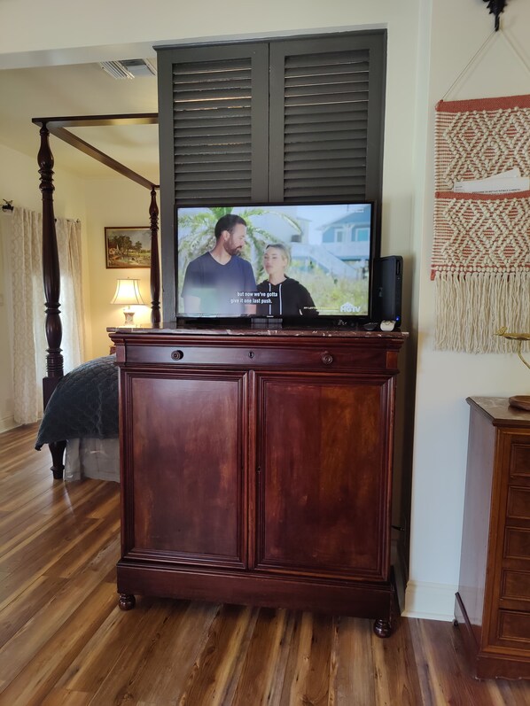 A television on a wooden cabinet with a dark background, a bed with a white bedspread, and a wall-mounted tapestry.