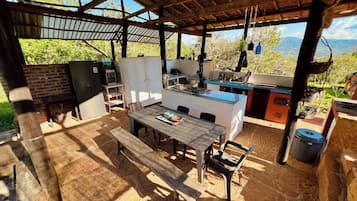 An outdoor kitchen area with a dining table and chairs, surrounded by stone flooring and a wooden pergola.
