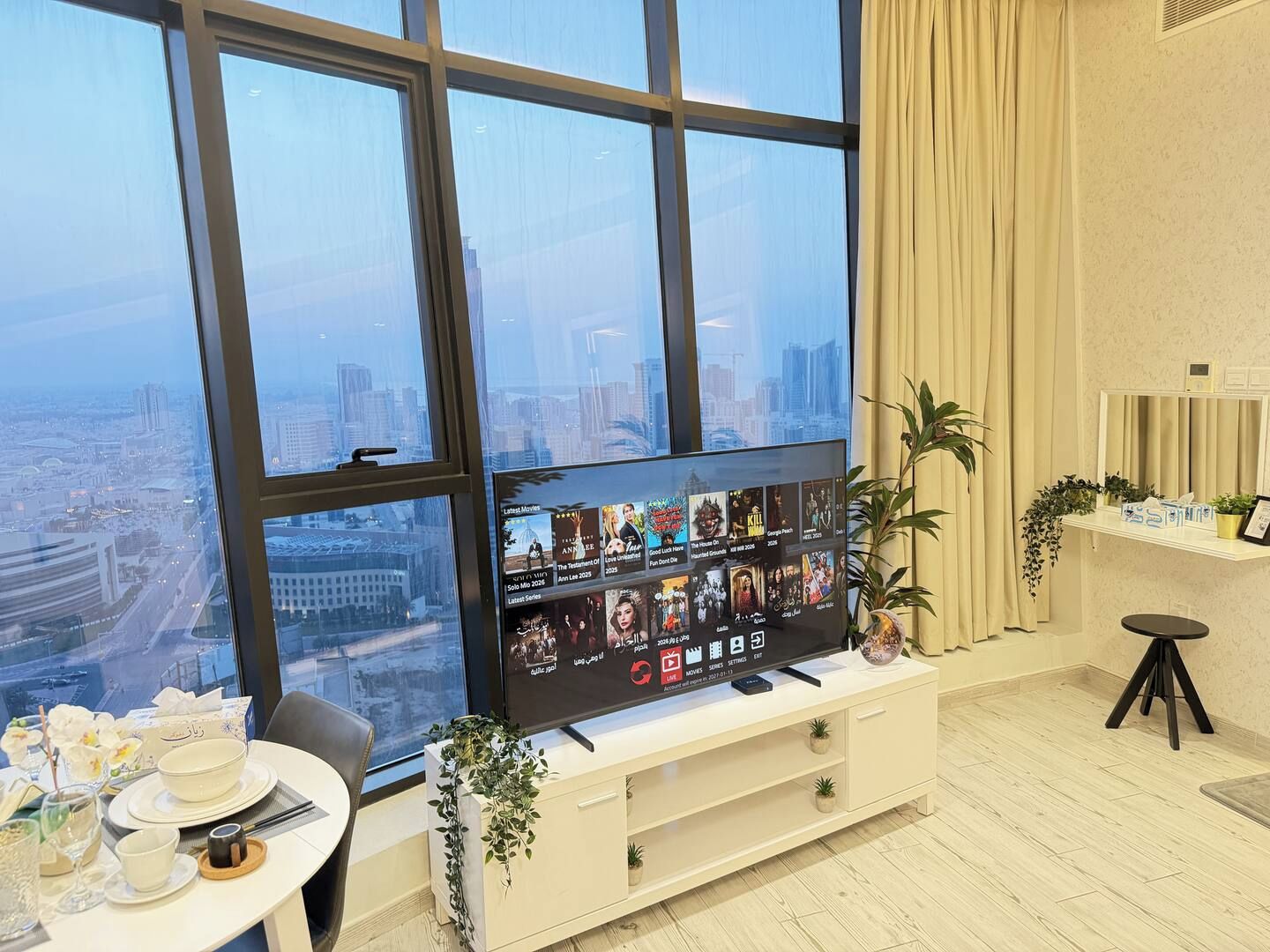 A modern room with a large window, a flat-screen TV, a dining table with a bowl and chopsticks, and a small stool.