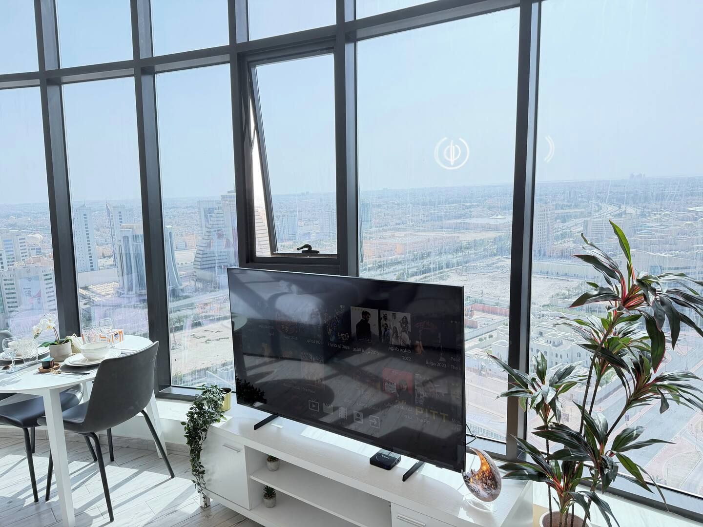 A modern living room with a large TV, a dining table, chairs, and a view of a cityscape through large windows.