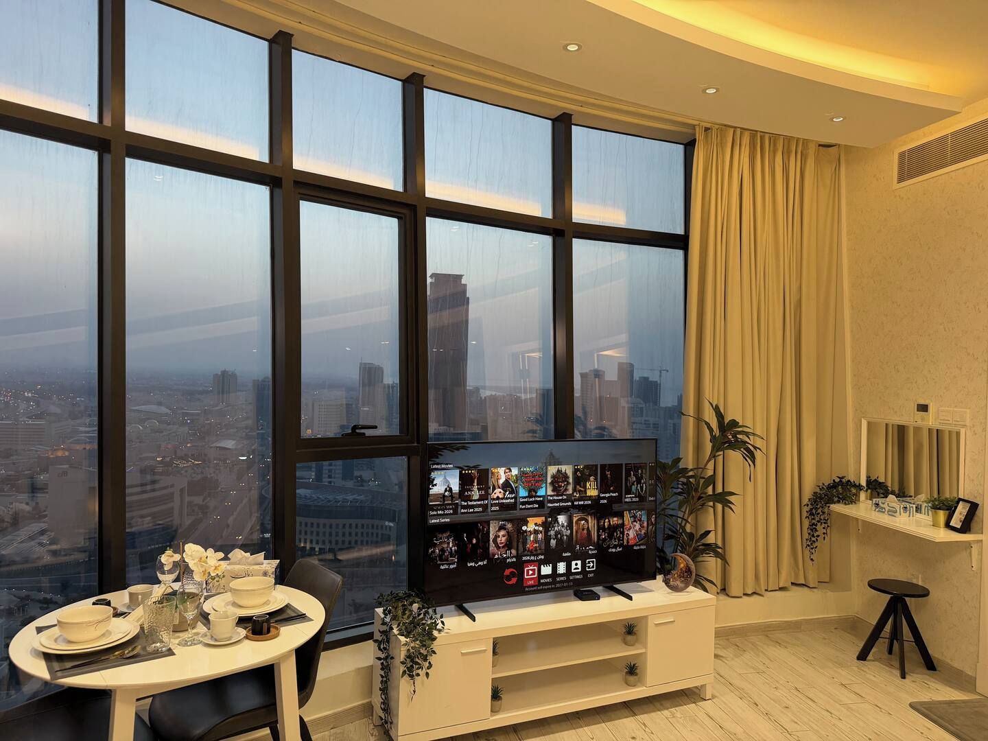 A modern living room with a large window, a flat-screen TV, a dining table with a vase of flowers, and a small stool.