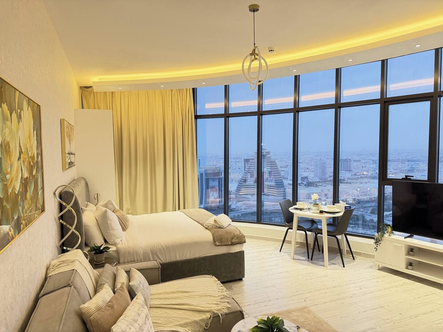 A modern hotel room with a large city view, a bed, a sofa, a dining table with chairs, and a TV.
