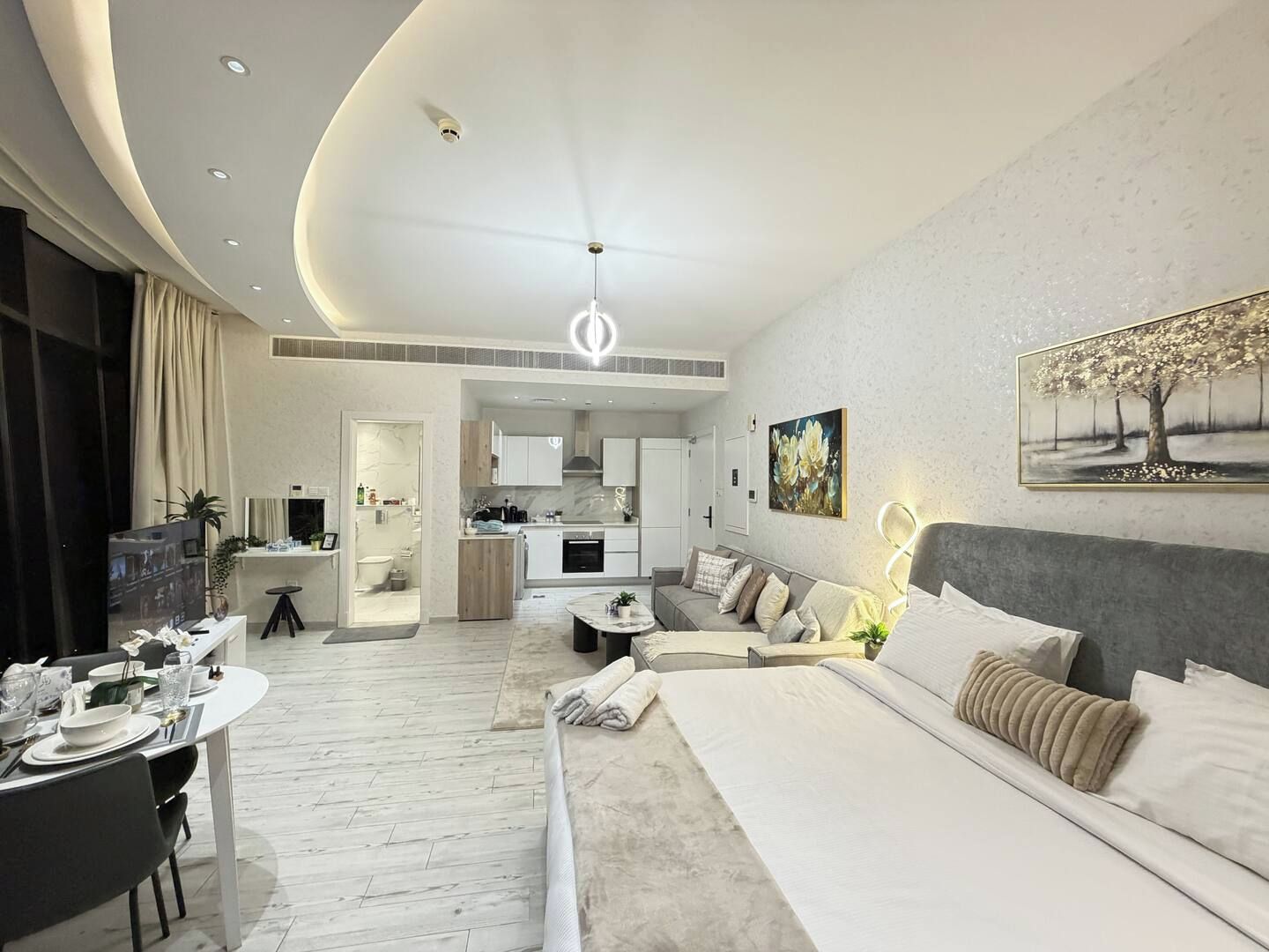 A modern hotel room with a curved ceiling, a dining area, a kitchen, and a living area with a sofa and a painting.