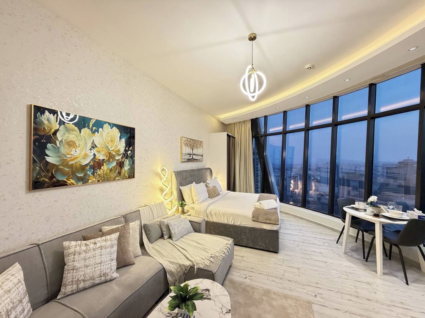 A modern hotel room with a large bed, a sofa, a dining table, and a view of the city through large windows.