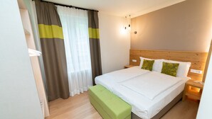 Hotellihuone, jossa on puinen sängynpääty, suuri sänky valkoisilla vuodevaatteilla, vihreä penkki ja ikkuna, jossa on verhot.