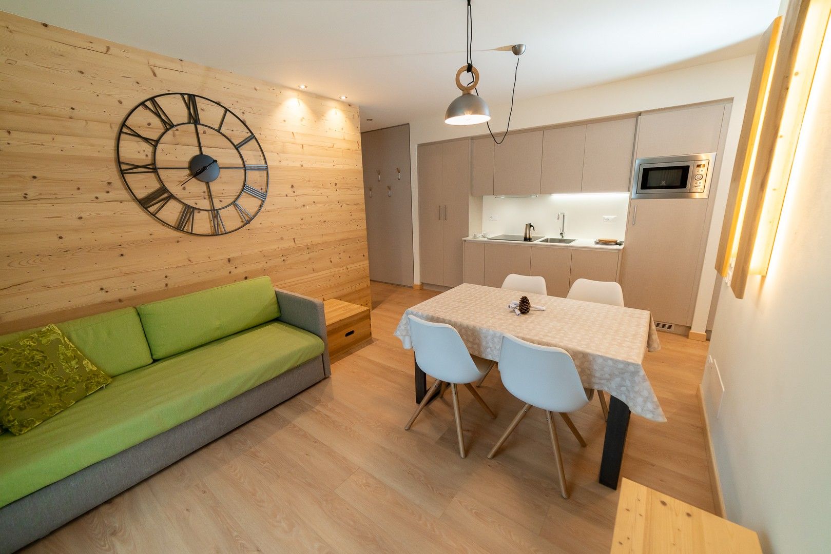 Een moderne woonruimte met een groene bank, een eettafel met stoelen, een grote wandklok en een keuken met een magnetron.