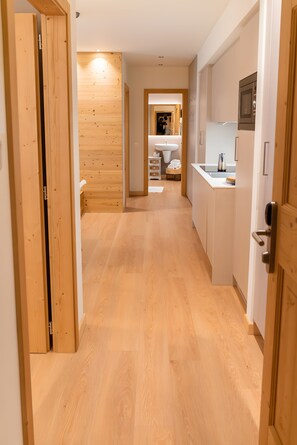 A hallway with wooden flooring leading to a kitchen area with white cabinets and a built-in microwave.