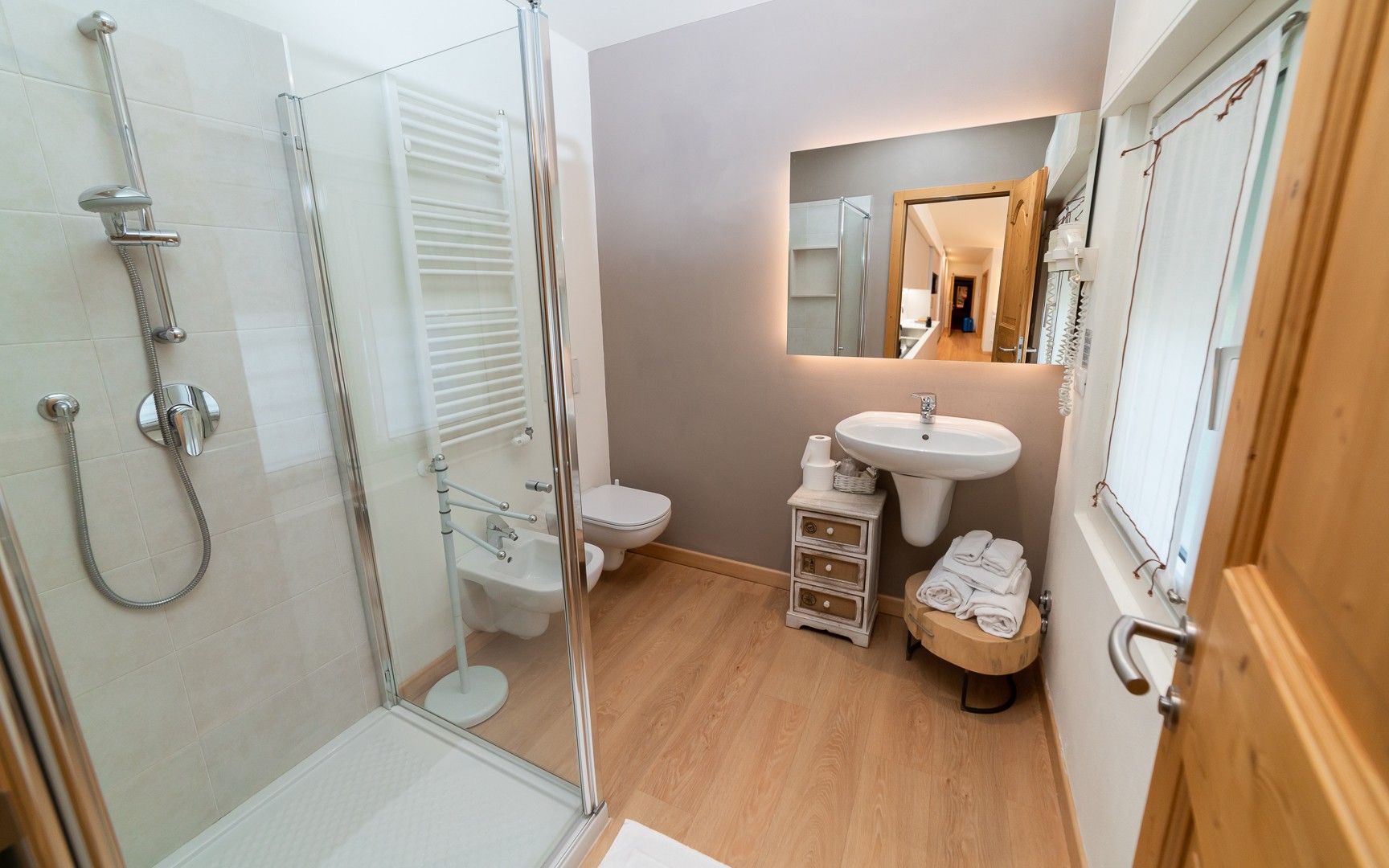 Une salle de bain moderne avec une douche à parois de verre, des toilettes blanches, un lavabo avec un miroir au-dessus et un petit tabouret en bois sur lequel sont posées des serviettes.