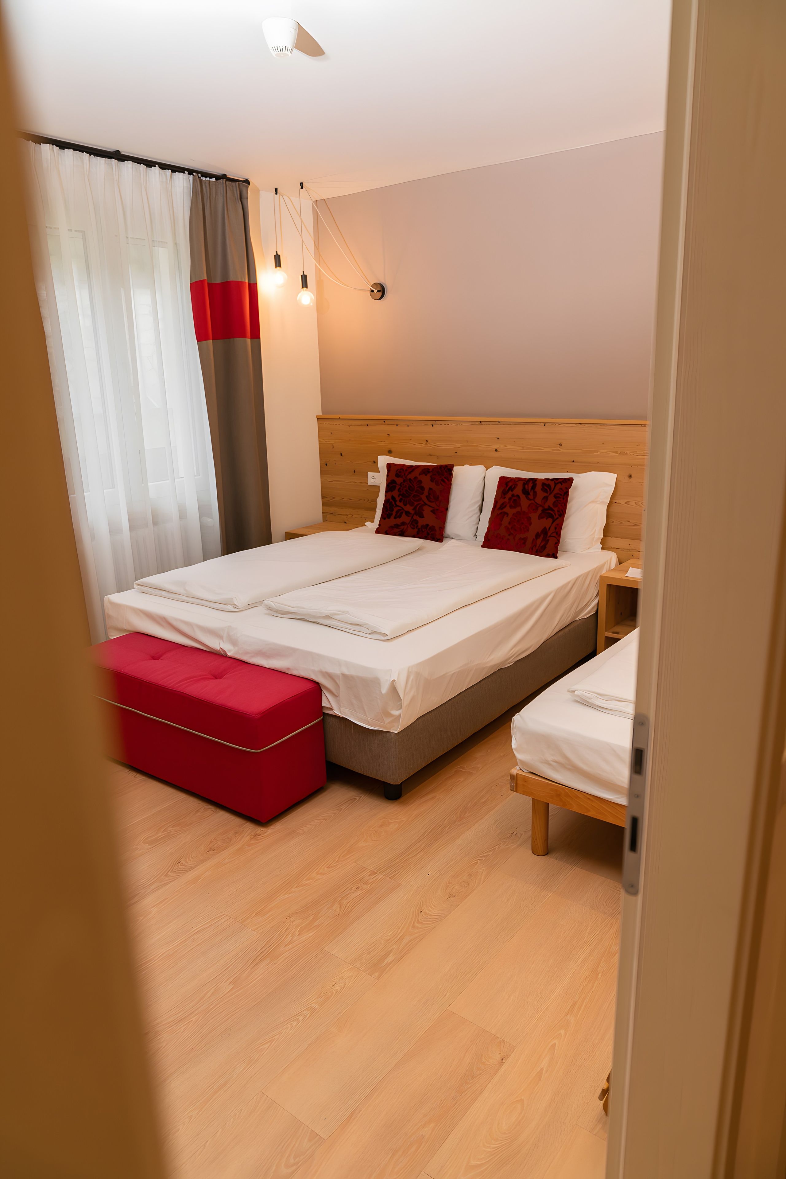 Un dormitorio con una cama doble, un reposapiés rojo, una mesita de noche de madera y una lámpara de pared.