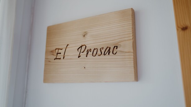 Una placa de madera con la inscripción 'El Prosac' fijada en una pared blanca.