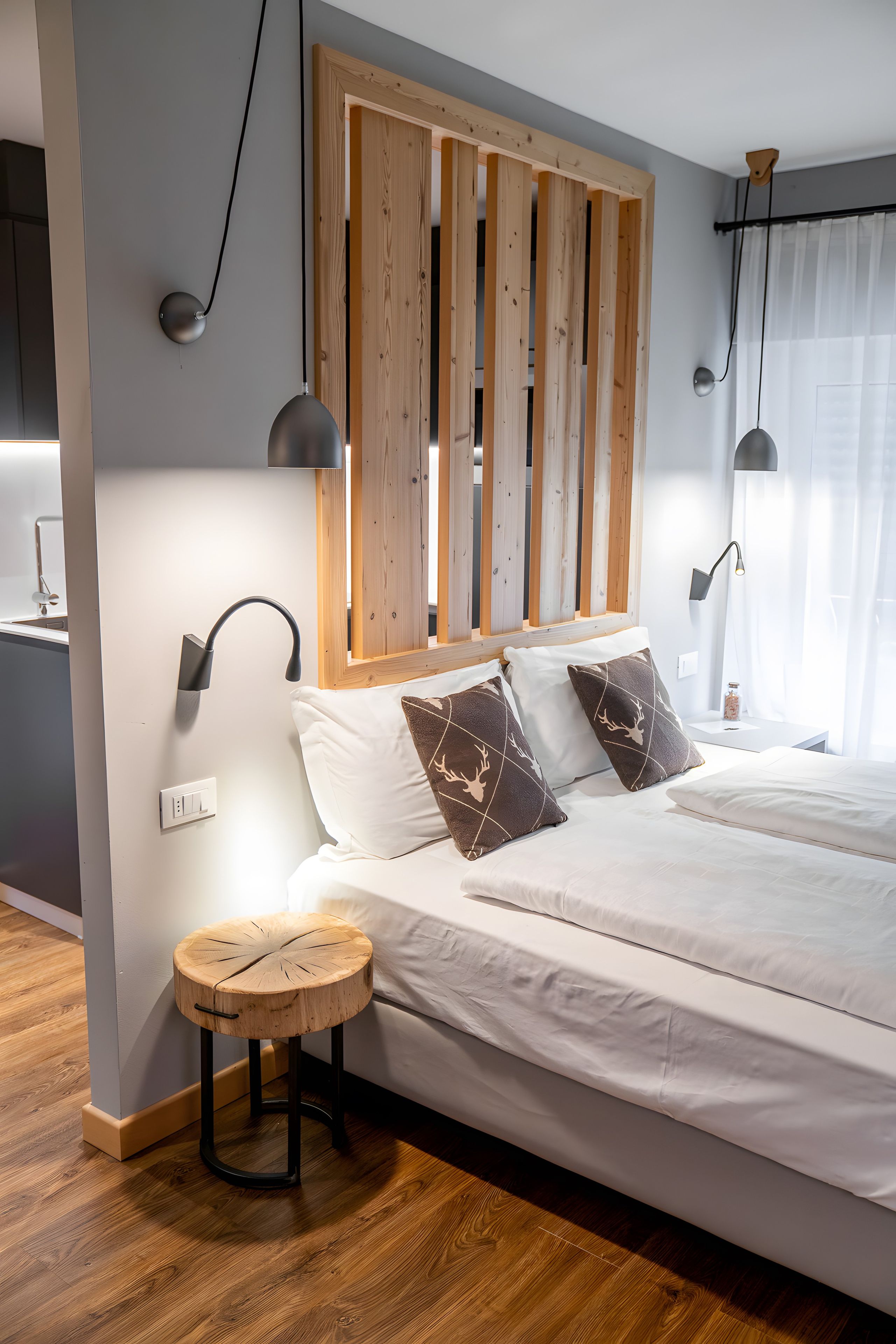 Een moderne slaapkamer met een houten hoofdbord, een rond houten krukje, een bed met wit beddengoed en gestreepte kussens, en twee wandlampen.