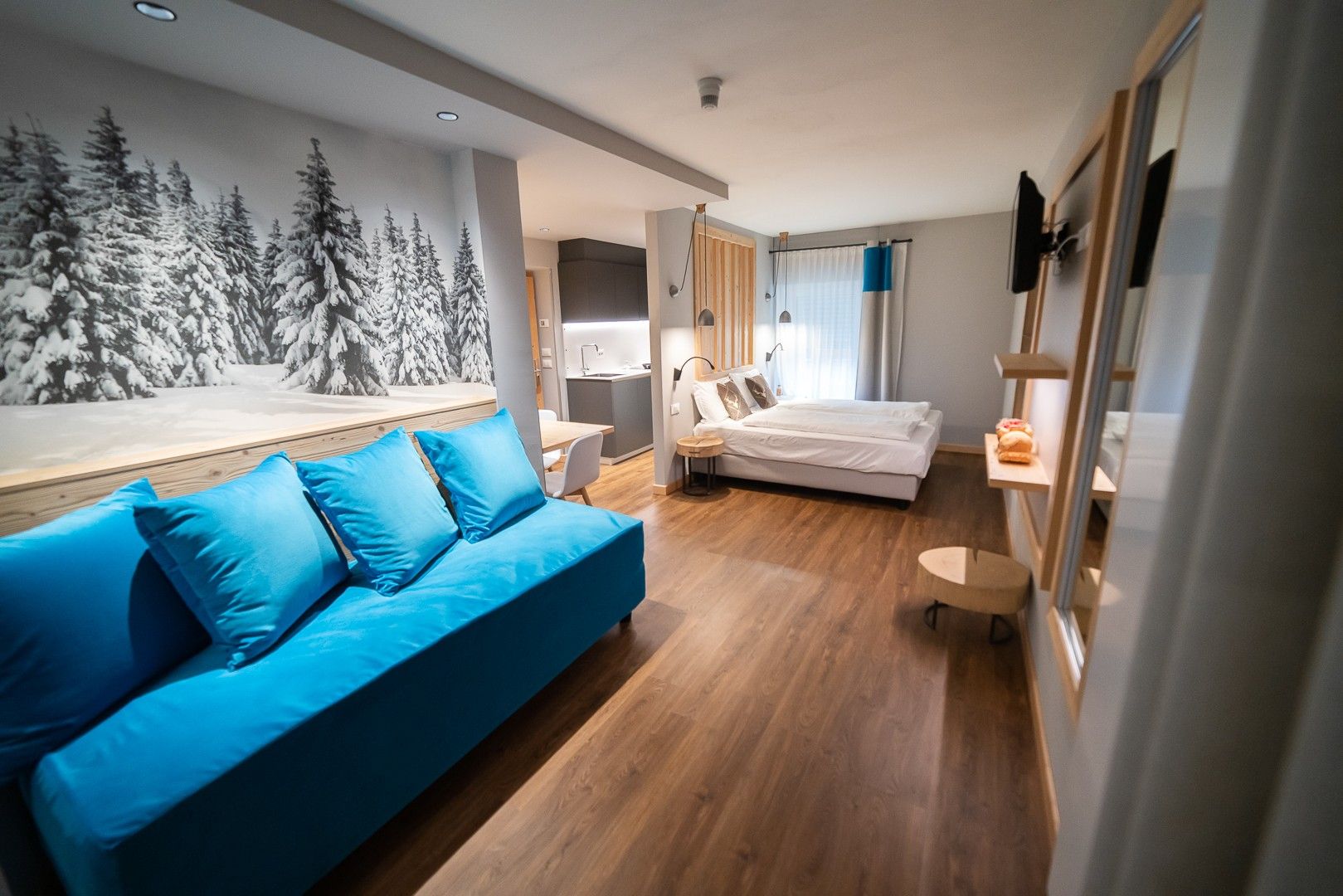 Une chambre moderne avec un canapé bleu, un lit, un bureau et un mur orné d’une fresque représentant une forêt.