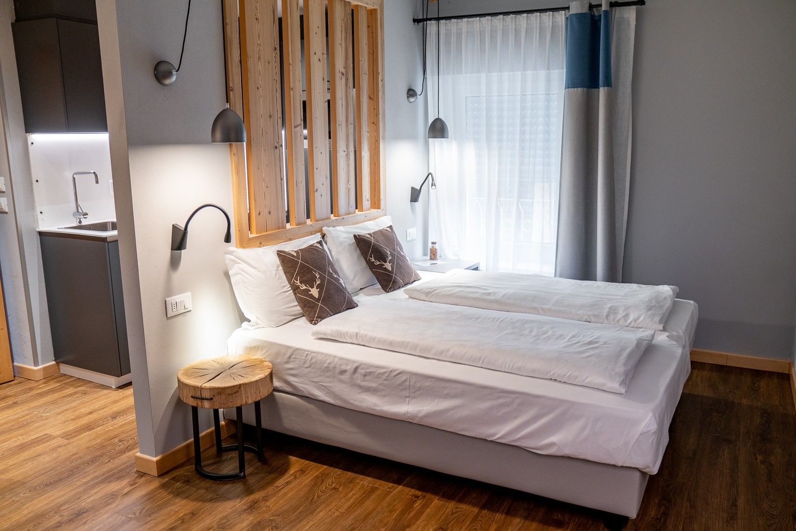Une chambre moderne avec un lit, des lampes de chevet, une tête de lit en bois, une petite table ronde et un coin cuisine intégré.