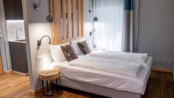 Une chambre moderne avec un lit, des lampes de chevet, une tête de lit en bois, une petite table ronde et un coin cuisine intégré.