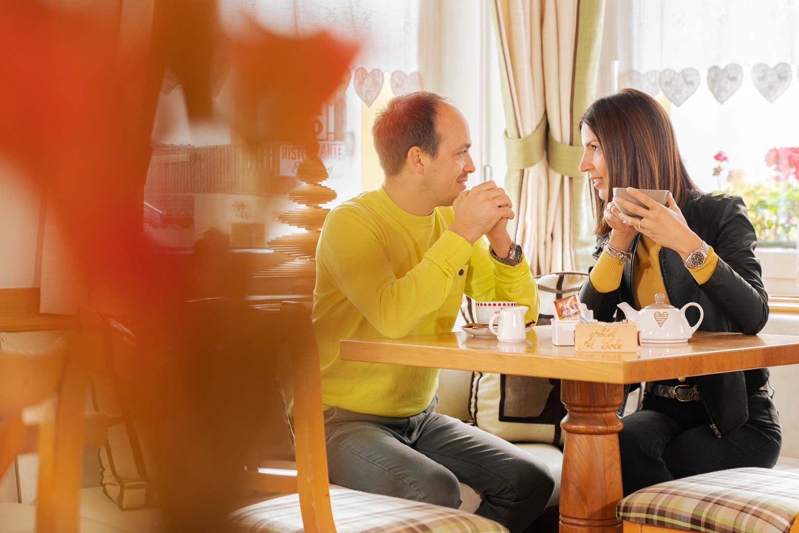 Twee personen zitten aan een houten tafel met een theepot en kopjes, in een kamer met gordijnen en decoraties in de vorm van harten.