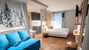 Habitación de hotel moderna con suelo de madera, sofá azul, cama, escritorio y televisor montado en la pared.