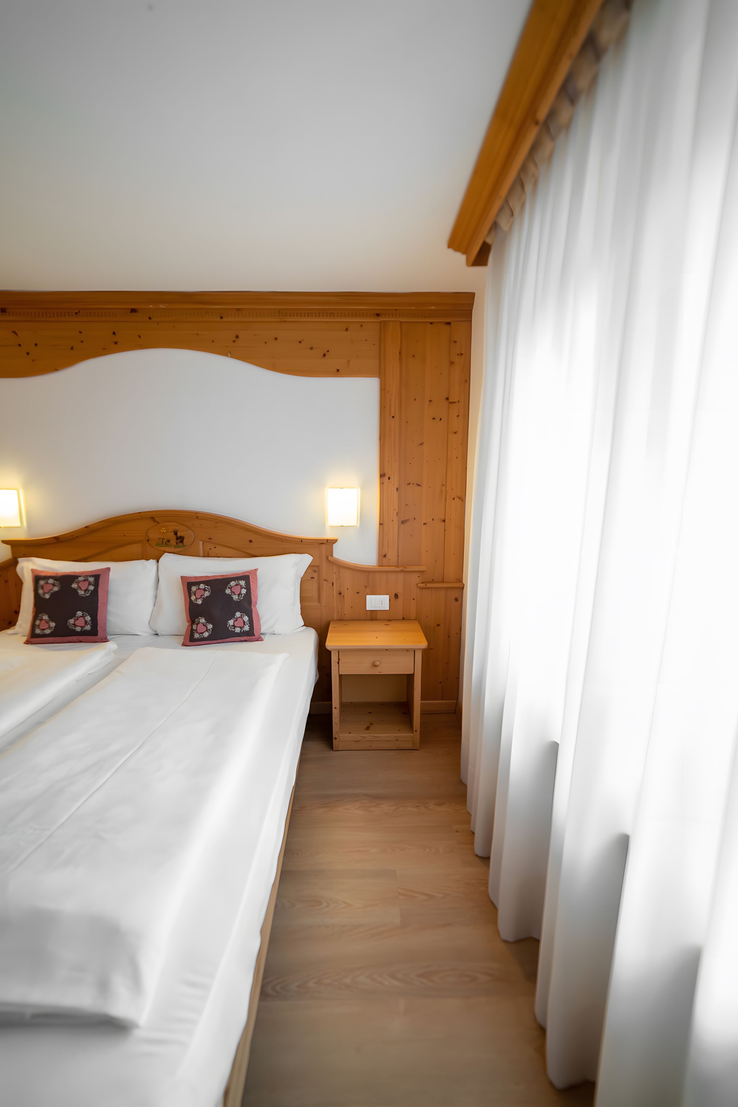 Ein Hotelzimmer mit Holz-Kopfteil, einem Bett mit weißer Bettwäsche, zwei Zierkissen, einem Nachttisch mit Lampe und einem Fenster mit Vorhängen.