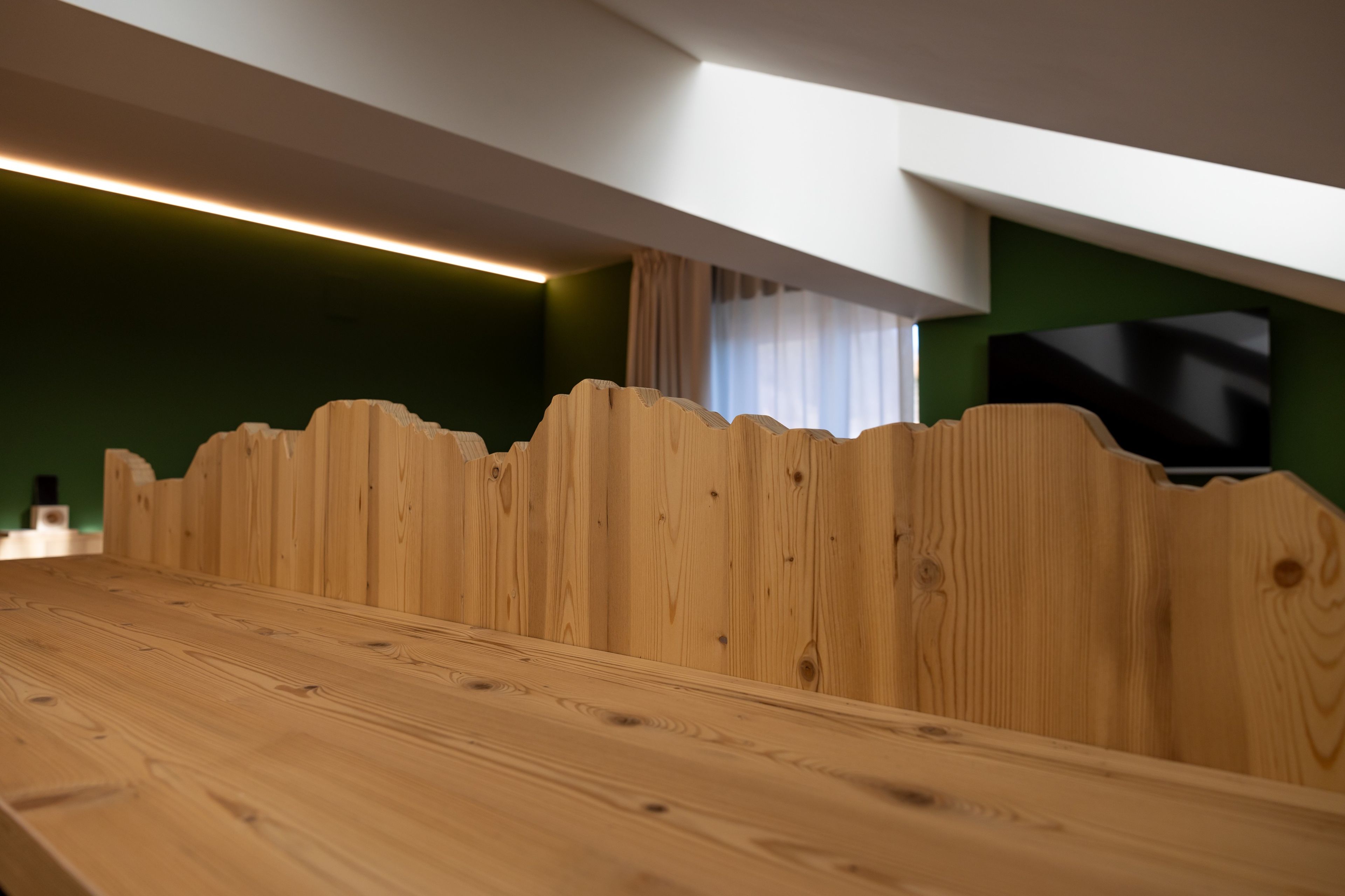 En träbänk med en unik, vågformad design på ryggstödet, en plan yta och en modern inredning med gröna väggar och en taklampa.