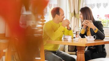 Two people seated at a wooden table with a teapot and cups, in a room with curtains and heart-shaped decorations.
