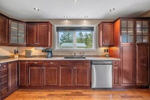 A modern kitchen with wooden cabinets, a stainless steel dishwasher, and a window with a view of trees.