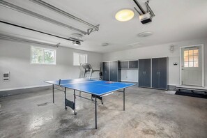 A room with a ping pong table, ceiling lights, windows, and cabinets.