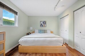 Un dormitorio con cama, mesita de noche, lámpara, una obra de arte y vistas a la vegetación por la ventana.