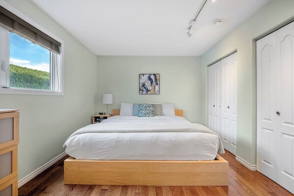 A bedroom with a bed, bedside table, lamp, artwork, and a view of greenery through the window.