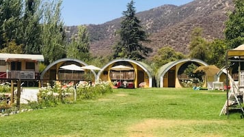 A series of dome-shaped buildings with outdoor seating areas, surrounded by a grassy field and trees, with a mountainous backdrop.
