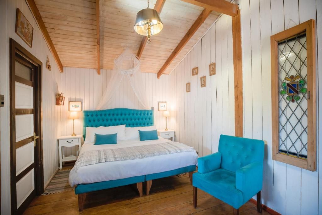 A bedroom with a bed, bedside lamps, a chair, a window with a decorative glass, and wooden beams on the ceiling.