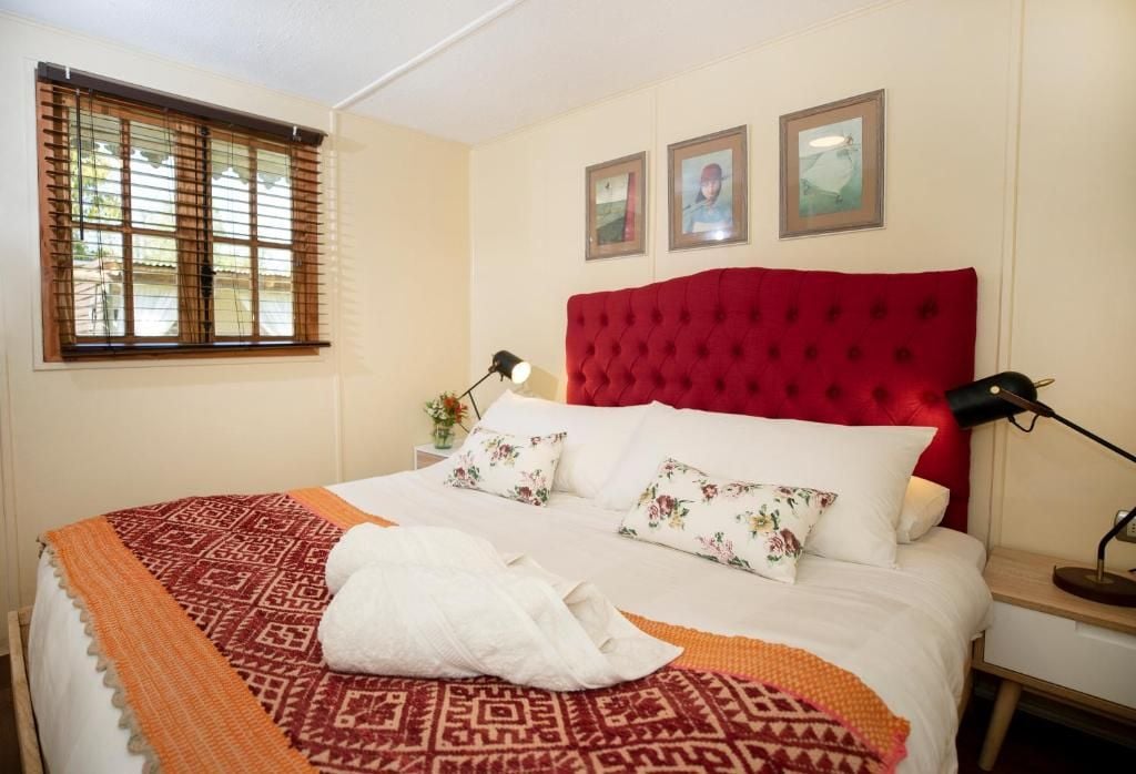 Une chambre d’hôtel avec un lit, une fenêtre avec des stores, une tête de lit rouge et trois tableaux encadrés accrochés au mur.