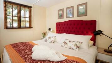 Une chambre d’hôtel avec un lit, une fenêtre avec des stores, une tête de lit rouge et trois tableaux encadrés accrochés au mur.