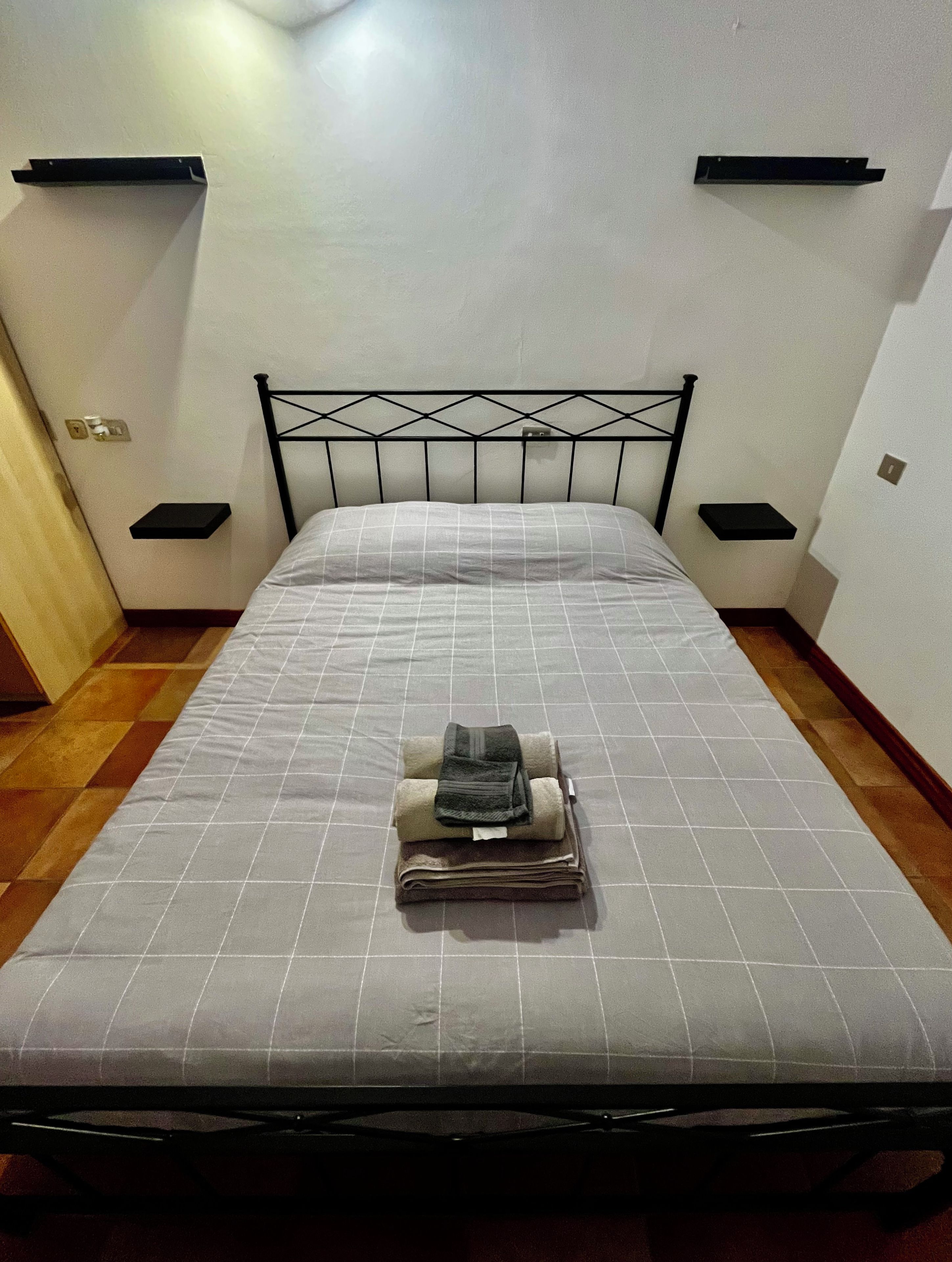 A bed with a metal frame, a grey mattress, and a folded towel on top. There are two bedside tables with black surfaces.