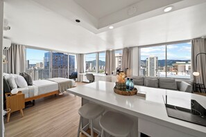 A modern hotel room with a large window, a sofa, a bed, a dining area, and a view of the cityscape. - 0.1 Mile to Beach 23FL 1BR 2BED Ocean/Mountain Views RF TOP Pool Central Waikiki (Honolulu)