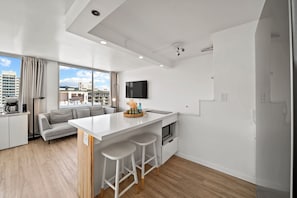A modern kitchen with a central island, bar stools, a flat-screen TV, and a view of the cityscape. - 0.1 Mile to Beach 23FL 1BR 2BED Ocean/Mountain Views RF TOP Pool Central Waikiki (Honolulu)