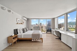 Ein modernes Schlafzimmer mit einem großen Bett, einem Schreibtisch und Blick auf eine Skyline durch raumhohe Fenster.
