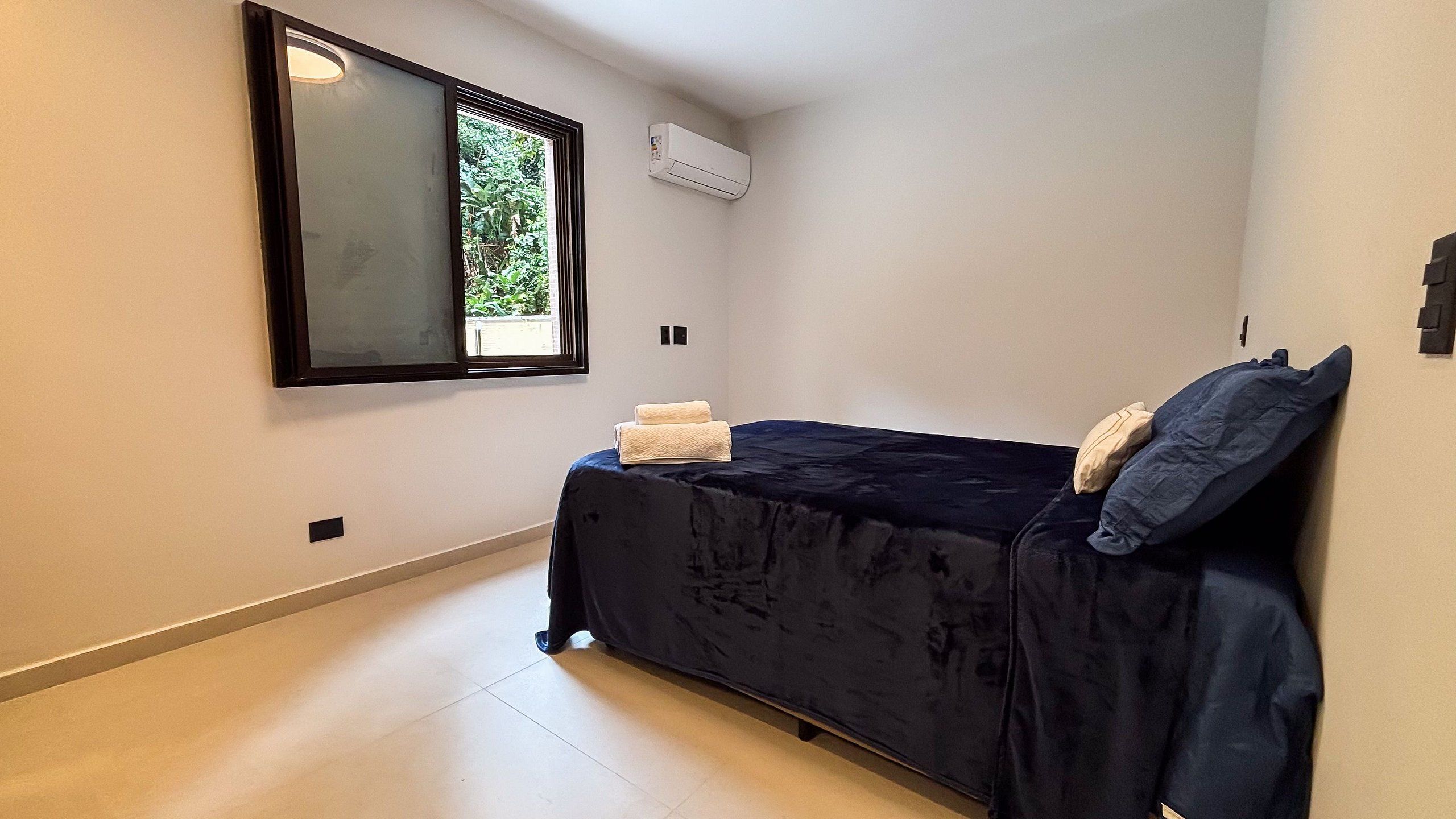 A small, single-bed room with a window, an air conditioning unit, and a tiled floor.