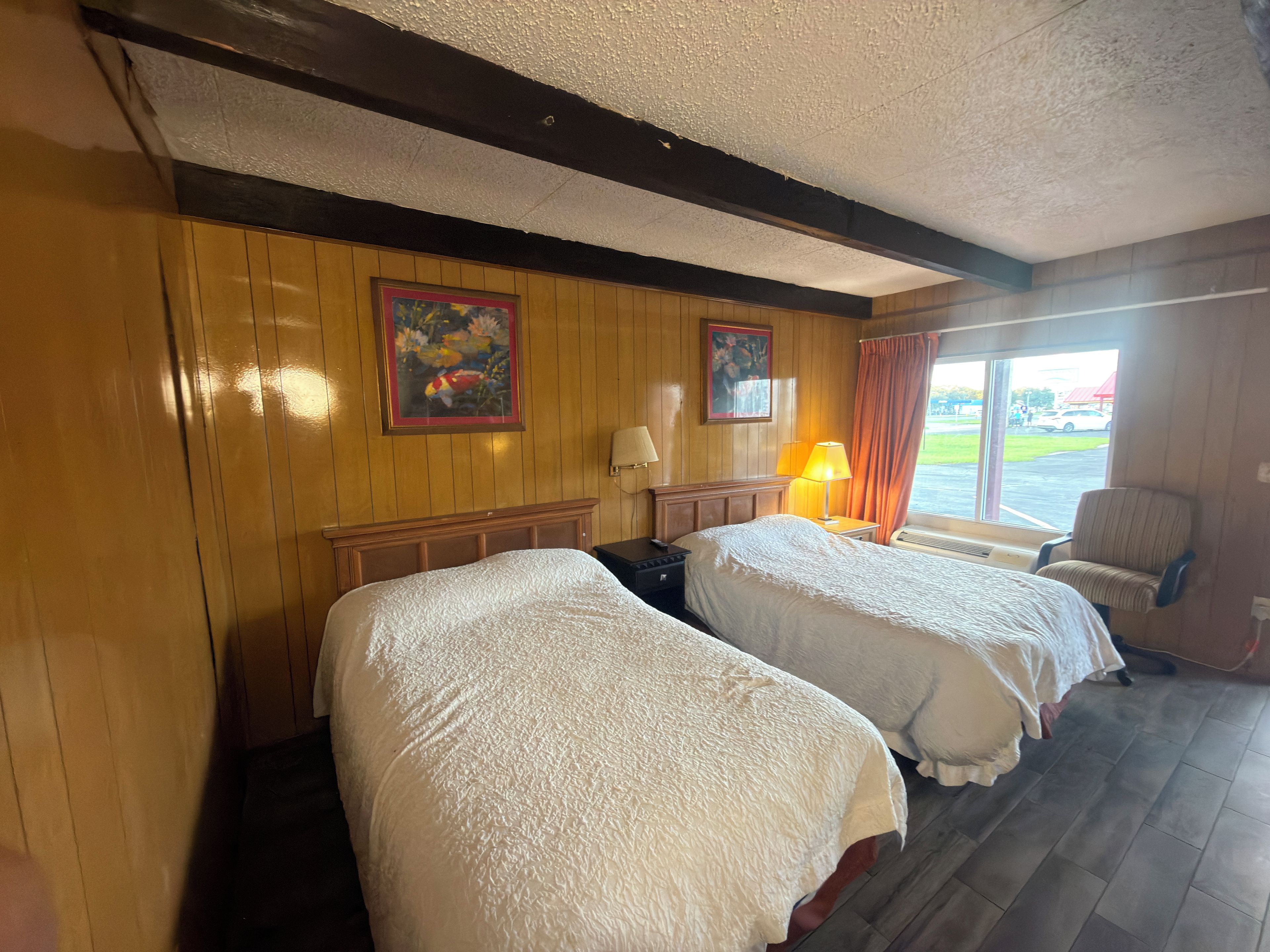 A hotel room with two beds, a wooden wall, a painting, a window with curtains, and a chair.