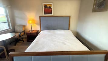 Uma cama de solteiro com cabeceira de madeira, mesa de cabeceira com abajur, uma cadeira e janela com vista para o exterior.