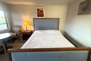 A single bed with a wooden headboard, a bedside table with a lamp, a chair, and a window with a view of the outdoors.