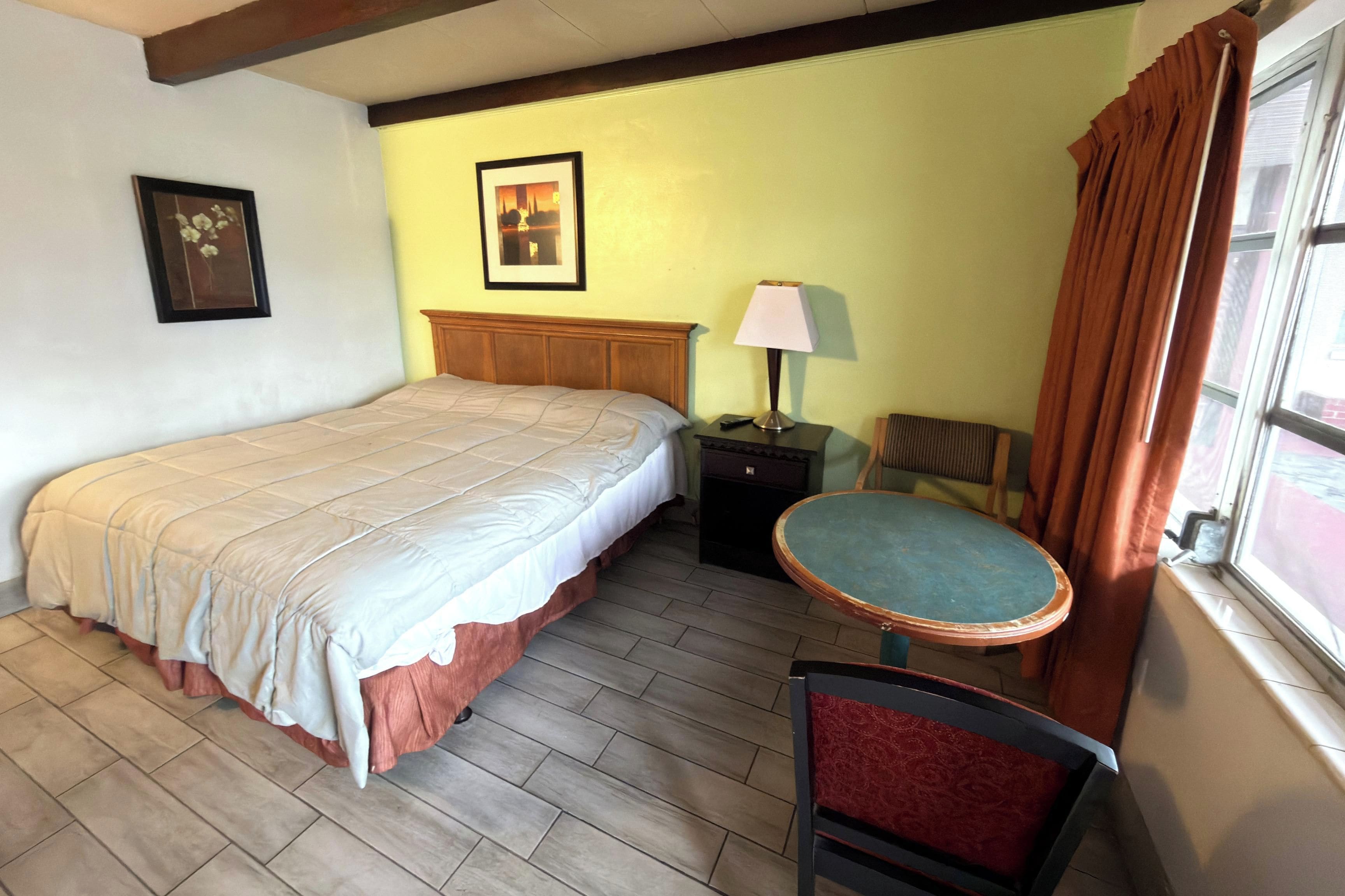 Une chambre d’hôtel avec un lit, une table de chevet, une lampe, une chaise et une fenêtre avec des rideaux.
