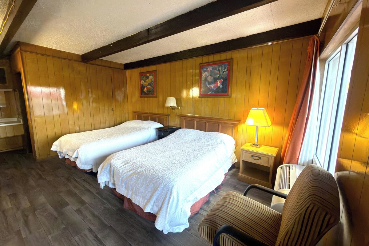 A room with two beds, a chair, a nightstand, a lamp, and a window with curtains.