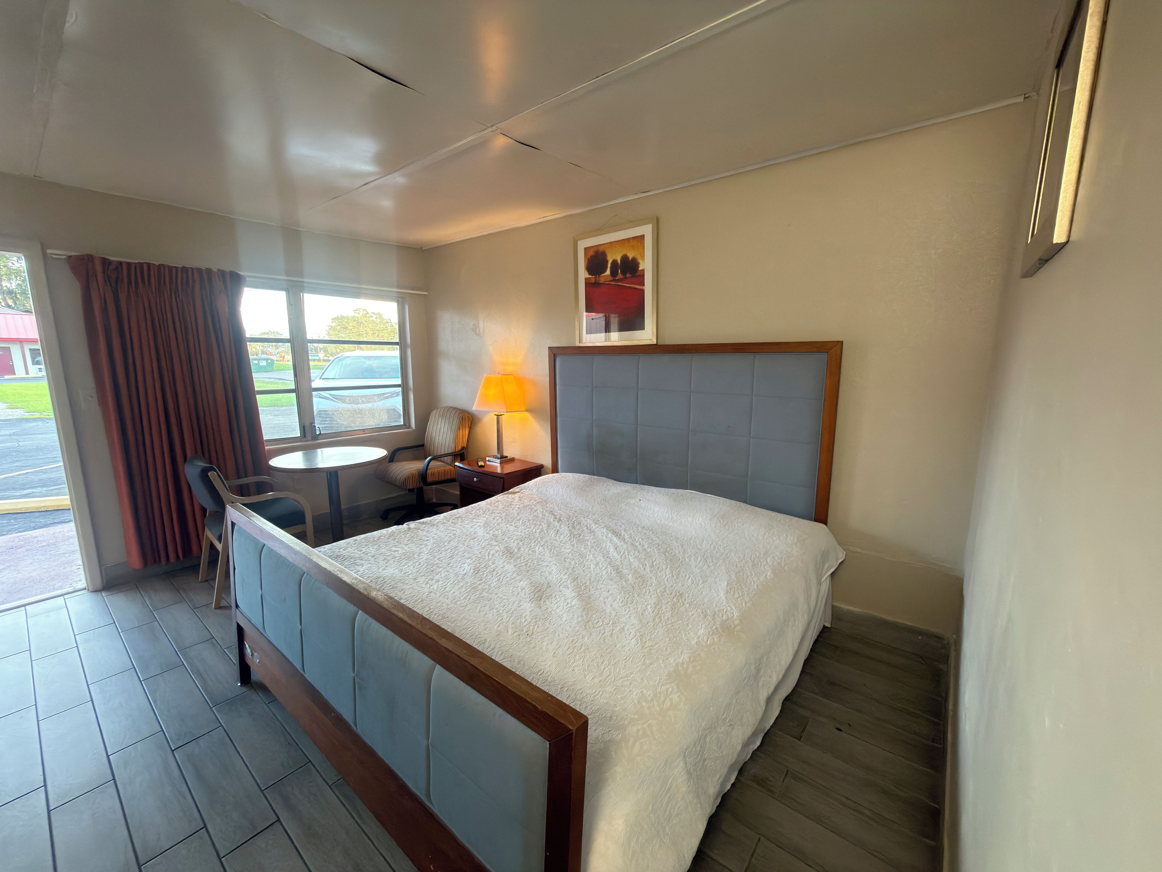 Ein Hotelzimmer mit einem Bett, einem Schreibtisch mit Stuhl, einer Lampe, einem Fenster mit Vorhängen und einem Bild an der Wand.